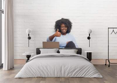 Young african american woman sitting on the floor using laptop at home doing happy thumbs up gesture with hand. Approving expression looking at the camera with showing success. Wall mural