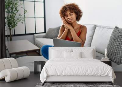 Young african american woman sitting on the sofa at home using laptop sleeping tired dreaming and posing with hands together while smiling with closed eyes. Wall mural