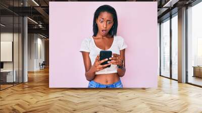 Young african american woman using smartphone in shock face, looking skeptical and sarcastic, surprised with open mouth Wall mural