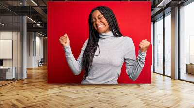 Young african american woman wearing turtleneck sweater over red isolated background very happy and excited doing winner gesture with arms raised, smiling and screaming for success. Celebration Wall mural