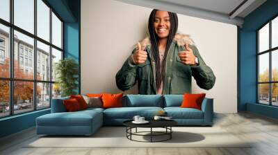 Young african american woman wearing winter parka coat over isolated background success sign doing positive gesture with hand, thumbs up smiling and happy. Cheerful expression and winner gesture. Wall mural