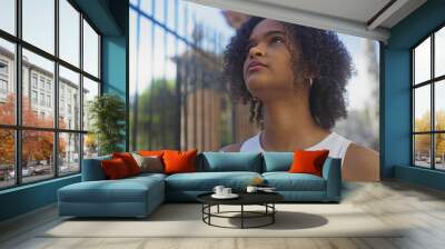 Young african american woman with curly hair looking up on an urban street during daytime. Wall mural