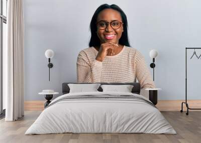 Young african woman wearing casual clothes sitting on the table smiling looking confident at the camera with crossed arms and hand on chin. thinking positive. Wall mural