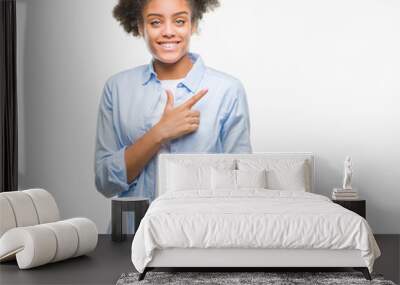 Young afro american woman over isolated background cheerful with a smile of face pointing with hand and finger up to the side with happy and natural expression on face looking at the camera. Wall mural