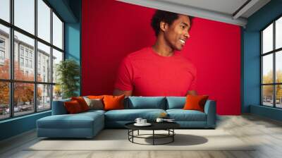 Young american man with afro hair wearing t-shirt standing over isolated red background looking away to side with smile on face, natural expression. Laughing confident. Wall mural