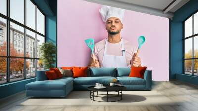 Young arab man wearing professional cook apron and hat holding spoon looking at the camera blowing a kiss being lovely and sexy. love expression. Wall mural