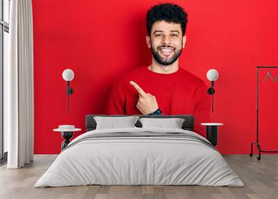 Young arab man with beard wearing casual red sweater cheerful with a smile of face pointing with hand and finger up to the side with happy and natural expression on face Wall mural