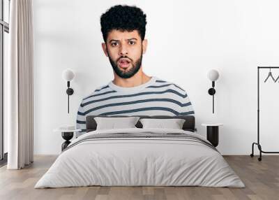 Young arab man with beard wearing casual striped sweater in shock face, looking skeptical and sarcastic, surprised with open mouth Wall mural