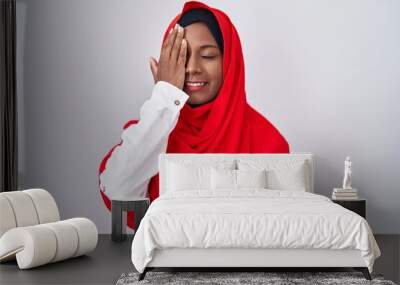 Young arab woman wearing traditional islamic hijab scarf covering one eye with hand, confident smile on face and surprise emotion. Wall mural