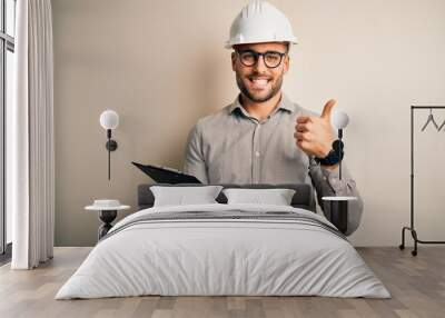 Young architect inspector man wearing builder safety helmet and using clipboard happy with big smile doing ok sign, thumb up with fingers, excellent sign Wall mural