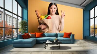 Young asian woman holding paper bag of fresh healthy groceries over yellow isolated background smiling with happy face winking at the camera doing victory sign with fingers. Number two. Wall mural