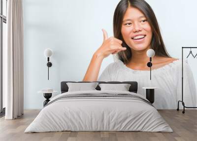 Young asian woman over isolated background smiling doing phone gesture with hand and fingers like talking on the telephone. Communicating concepts. Wall mural