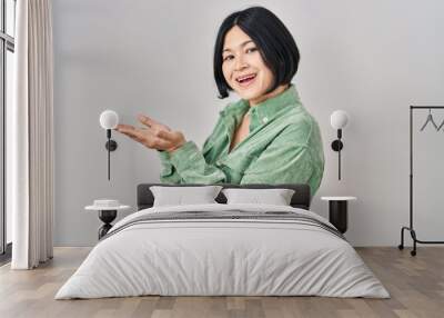 Young asian woman standing over white background pointing aside with hands open palms showing copy space, presenting advertisement smiling excited happy Wall mural