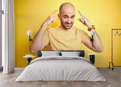 Young bald man with beard wearing casual striped t-shirt over yellow isolated background smiling pointing to head with both hands finger, great idea or thought, good memory Wall mural