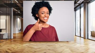 Young beautiful African American afro woman with curly hair wearing casual t-shirt standing doing happy thumbs up gesture with hand. Approving expression looking at the camera showing success. Wall mural