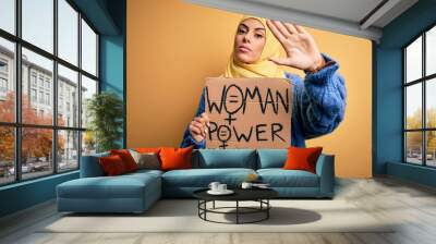 Young beautiful arab woman wearing islamic hijab holding banner with women power message with open hand doing stop sign with serious and confident expression, defense gesture Wall mural