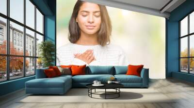 Young beautiful arab woman wearing winter sweater over isolated background smiling with hands on chest with closed eyes and grateful gesture on face. Health concept. Wall mural