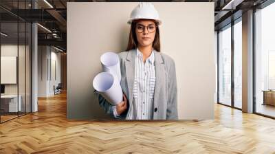 Young beautiful architect woman wearing safety helmet and glasses holding blueprints with a confident expression on smart face thinking serious Wall mural