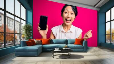 Young beautiful asian girl holding smartphone showing screen over isolated pink background screaming proud and celebrating victory and success very excited, cheering emotion Wall mural