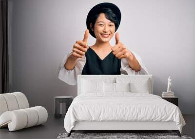 Young beautiful asian girl wearing casual dress and hat standing over isolated white background approving doing positive gesture with hand, thumbs up smiling and happy for success. Winner gesture. Wall mural