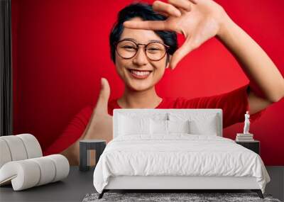 Young beautiful asian girl wearing casual t-shirt and glasses over isolated red background smiling making frame with hands and fingers with happy face. Creativity and photography concept. Wall mural