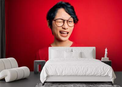 Young beautiful asian girl wearing casual t-shirt and glasses over isolated red background winking looking at the camera with sexy expression, cheerful and happy face. Wall mural