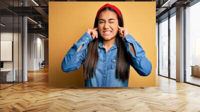 Young beautiful asian woman wearing casual denim shirt and diadem over yellow background covering ears with fingers with annoyed expression for the noise of loud music. Deaf concept. Wall mural