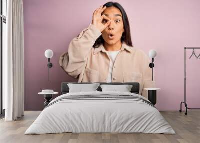 Young beautiful asian woman wearing casual shirt standing over pink background doing ok gesture shocked with surprised face, eye looking through fingers. Unbelieving expression. Wall mural