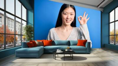 Young beautiful asian woman wearing casual sweater standing over blue isolated background smiling positive doing ok sign with hand and fingers. Successful expression. Wall mural