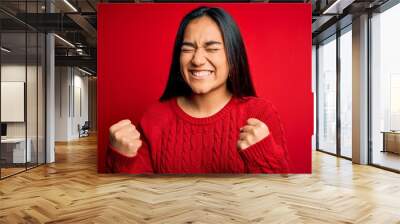 Young beautiful asian woman wearing casual sweater standing over isolated red background very happy and excited doing winner gesture with arms raised, smiling and screaming for success. Celebration Wall mural