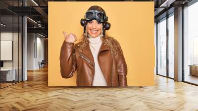 Young beautiful blonde aviator woman wearing vintage pilot helmet whit glasses and jacket smiling with happy face looking and pointing to the side with thumb up. Wall mural