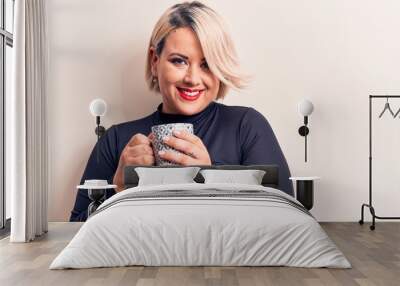 Young beautiful blonde plus size woman drinking mug of coffee over white background looking positive and happy standing and smiling with a confident smile showing teeth Wall mural