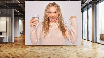 Young beautiful blonde woman drinking glass of water over isolated white background screaming proud, celebrating victory and success very excited with raised arms Wall mural