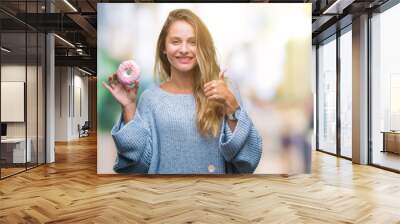 Young beautiful blonde woman eating sweet donut over isolated background happy with big smile doing ok sign, thumb up with fingers, excellent sign Wall mural