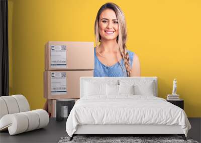 Young beautiful blonde woman holding delivery boxes looking positive and happy standing and smiling with a confident smile showing teeth Wall mural