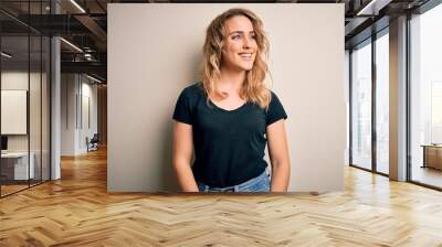 Young beautiful blonde woman wearing casual t-shirt standing over isolated white background looking away to side with smile on face, natural expression. Laughing confident. Wall mural