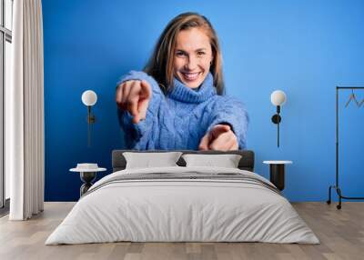 Young beautiful blonde woman wearing casual turtleneck sweater over blue background pointing to you and the camera with fingers, smiling positive and cheerful Wall mural