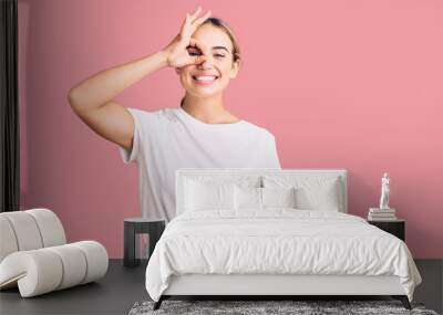 Young beautiful blonde woman wearing casual white tshirt doing ok gesture with hand smiling, eye looking through fingers with happy face. Wall mural