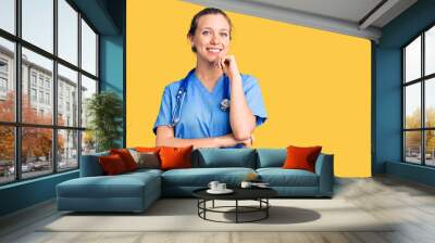 Young beautiful blonde woman wearing doctor uniform and stethoscope looking confident at the camera with smile with crossed arms and hand raised on chin. thinking positive. Wall mural