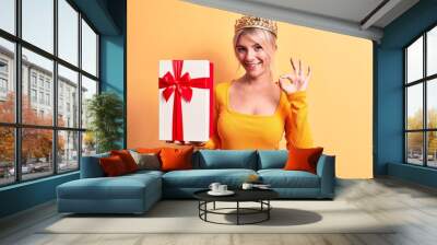 Young beautiful blonde woman wearing golden princess crown holding birthday gift doing ok sign with fingers, smiling friendly gesturing excellent symbol Wall mural