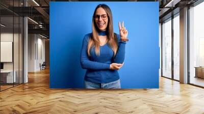 Young beautiful blonde woman with blue eyes wearing glasses standing over blue background smiling with happy face winking at the camera doing victory sign. Number two. Wall mural