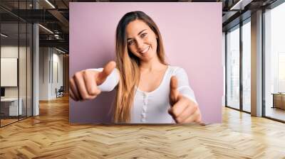 Young beautiful blonde woman with blue eyes wearing white t-shirt over pink background approving doing positive gesture with hand, thumbs up smiling and happy for success. Winner gesture. Wall mural