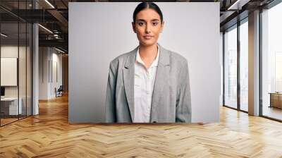 Young beautiful brunette businesswoman wearing jacket standing over white background with serious expression on face. Simple and natural looking at the camera. Wall mural