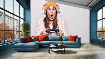 Young beautiful brunette gamer woman playing video game using joystick and headphones scared and amazed with open mouth for surprise, disbelief face Wall mural