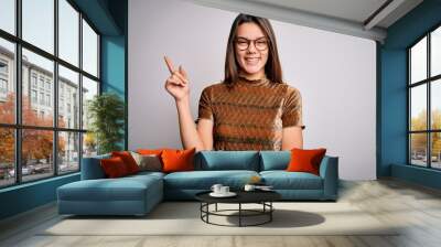 Young beautiful brunette girl wearing casual t-shirt and glasses over isolated white background with a big smile on face, pointing with hand and finger to the side looking at the camera. Wall mural