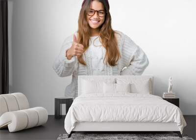 Young beautiful brunette hipster woman wearing glasses and winter hat over isolated background doing happy thumbs up gesture with hand. Approving expression looking at the camera showing success. Wall mural