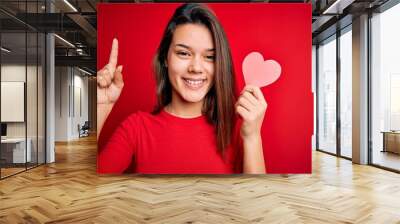 Young beautiful brunette romantic girl holding red paper heart shape over isolated background surprised with an idea or question pointing finger with happy face, number one Wall mural