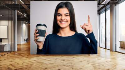 Young beautiful brunette woman drinking cup of coffee over isolated white background surprised with an idea or question pointing finger with happy face, number one Wall mural