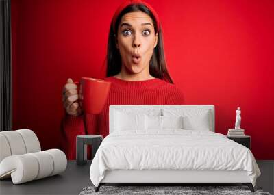 Young beautiful brunette woman drinking mug of coffe over isolated red background scared in shock with a surprise face, afraid and excited with fear expression Wall mural