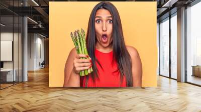 Young beautiful brunette woman holding asparagus scared and amazed with open mouth for surprise, disbelief face Wall mural